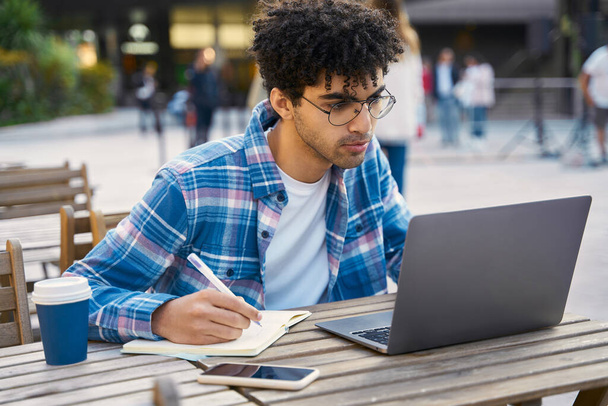 Fiatal gondolkodó férfi szabadúszó laptoppal, jegyzetel, tanfolyamokat néz a munkahelyén. Közel-keleti diák tanul, nyelvtanulás, online oktatási koncepció  - Fotó, kép