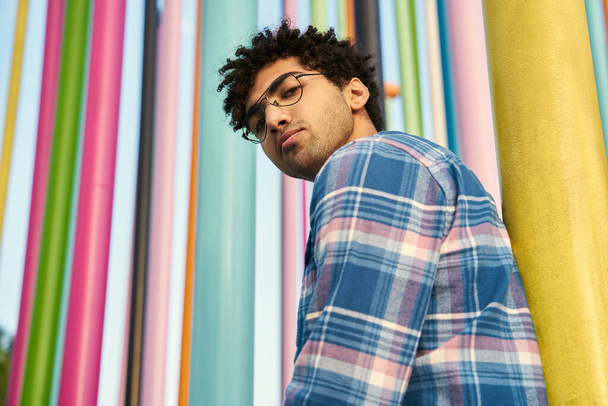 Low angle of the pensive middle eastern man wearing eyeglasses looking at the camera standing near the colorful wall. Human lifestyle concept  - Foto, Imagen