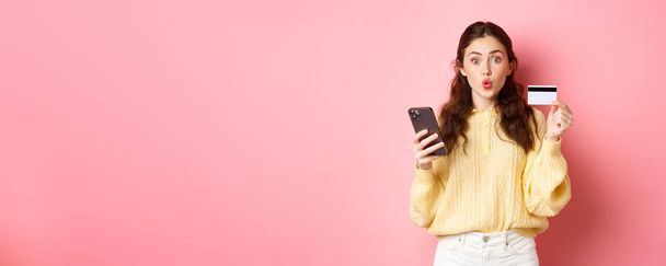 Технології та інтернет-магазини. Захоплений дівчина покупця, що показує пластикову кредитну картку і тримає мобільний телефон, платить зі смартфоном, замовляє онлайн, стоїть на рожевому фоні
. - Фото, зображення
