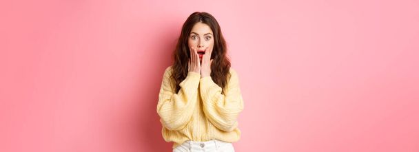 Image of shocked and surprised brunette woman gasping, checking out big news, open mouth, holding hands on face, standing amazed against pink background. - Zdjęcie, obraz