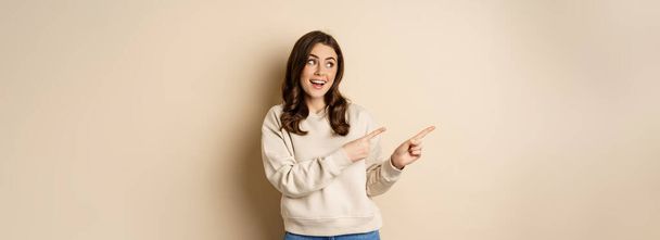 店の広告、バナーやロゴを表示し、左指を指す陽気魅力的な女性モデルは、ベージュの背景にセーターに立って. - 写真・画像