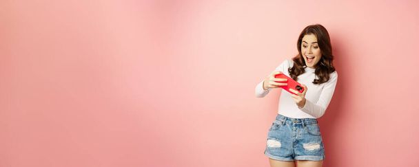 Happy beautiful girl playing mobile video game, holding smartphone horizontally, watching on cellphone with excited face, pink background. - Foto, immagini