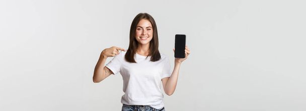 Ελκυστική χαμογελαστή γυναίκα δείχνοντας το δάχτυλο στην οθόνη smartphone, λευκό φόντο. - Φωτογραφία, εικόνα