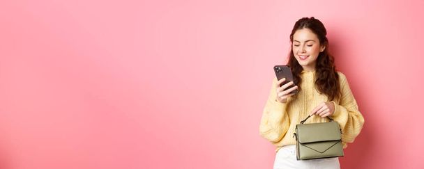 Glamour lány gazdaság pénztárca és szöveges üzenet okostelefon, olvasás mobiltelefon képernyő gondtalan mosollyal, álló felett rózsaszín háttér. - Fotó, kép