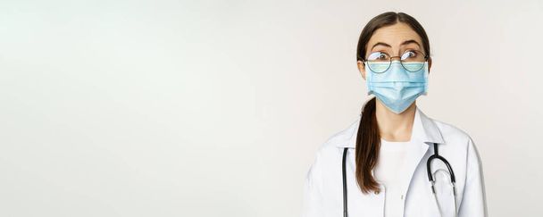 Medical worker, young doctor looking amazed and surprised, wearing face mask to prevent catching covid-19, standing over white background. - Foto, afbeelding