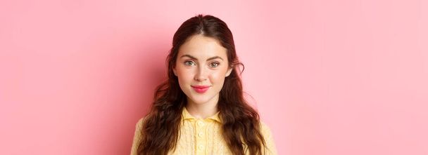 Close up of young 20s female model with cute feminine make up, smiling and looking hopeful at camera, standing against pink background. - Photo, Image