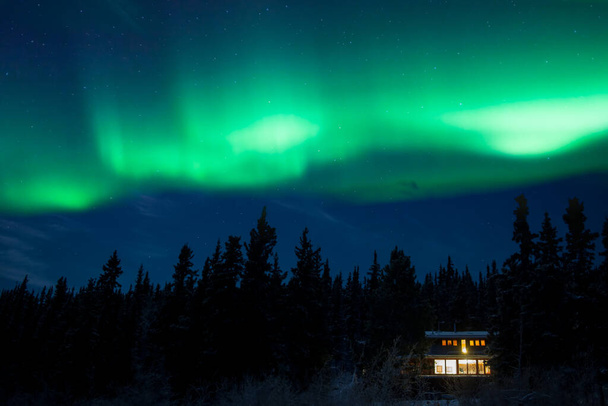 Cozy σπίτι taiga φωτίζεται θερμά κάτω από έναστρο νυχτερινό ουρανό με χορό βόρεια φώτα, Βόρειο Aurora, στο αρκτικό δάσος της Yukon Επικράτειας, Καναδάς - Φωτογραφία, εικόνα