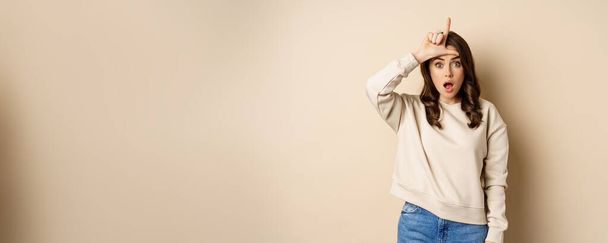 Woman showing loser sign on forehead, L word, standing over beige background. Copy space - Фото, изображение