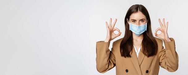 Workaplce and pandemic concept. Close up portrait of business woman showing okay signs, wearing medical mask, recommend, praise something, standing over white background. - Foto, afbeelding