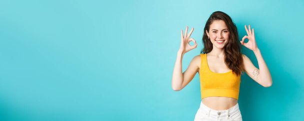 Beautiful and stylish confident woman showing okay signs, approve good choice, wearing summer clothes, standing over blue background. - Photo, Image