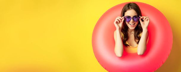 Close up portrait of stylish young woman, girl with swimming ring and summer sunglasses, ready for beach holiday, vacation, standing over yellow background. - Foto, Bild