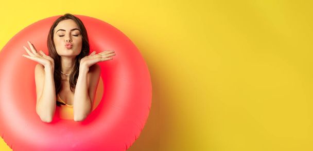 Beautiful coquettish woman inside pink swimming ring, posing on summer vacation, beach holiday concept, standing against yellow background. - Photo, Image