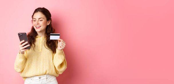 Tecnología y compras en línea. Joven linda dama que paga en línea con tarjeta de crédito, mirando el teléfono inteligente y sonriendo, de pie sobre el fondo rosa. - Foto, Imagen