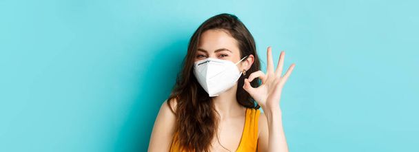 Covid-19, Coronavirus und soziale Distanzierung. Junge positive Frau in Atemschutzmaske zeigt Okay-Zeichen, steht vor blauem Hintergrund. - Foto, Bild