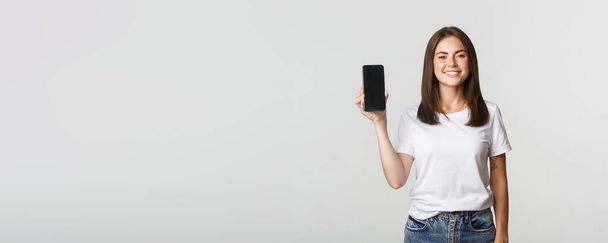 Attraktiv lächelnde junge Frau sieht zufrieden aus und zeigt Smartphone-Bildschirm. - Foto, Bild