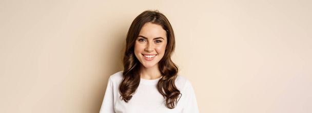 People. Close up portrait of young woman smiling, looking happy, wearing casual white t-shirt, standing healthy and cheerful against beige background. - Photo, image