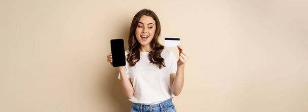 Online αγορές και οι άνθρωποι έννοια. Νεαρή όμορφη γυναίκα που αναζητούν ευτυχισμένη, δείχνει έκπτωση πιστωτικών καρτών και οθόνη του κινητού τηλεφώνου, στέκεται πάνω από το μπεζ φόντο. - Φωτογραφία, εικόνα