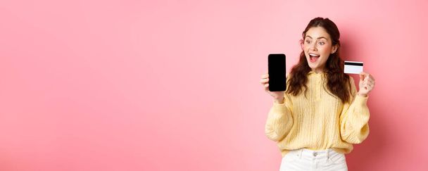 Technologie und Online-Shopping. Aufgeregtes attraktives Mädchen zeigt Smartphone-Bildschirm, Plastikkreditkarte, schaut erstaunt auf Telefon, steht vor rosa Hintergrund. - Foto, Bild