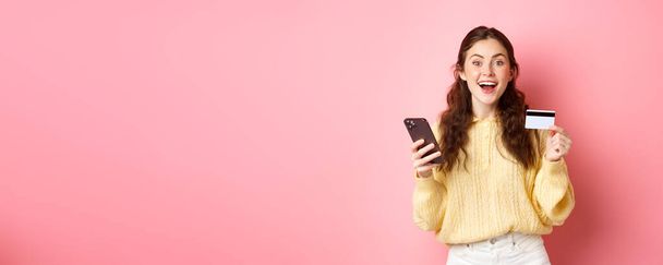 Τεχνολογία και online αγορές. Ενθουσιασμένη κοπέλα κάνοντας την παραγγελία, πληρώνοντας online με πλαστική πιστωτική κάρτα, κρατώντας το κινητό τηλέφωνο και χαμογελώντας στην κάμερα, ροζ φόντο. - Φωτογραφία, εικόνα