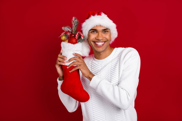 Photo of young toothy beaming smiling guy wear saint nicholas cap hold sock with adornment baubles xmas preparation isolated on red color background. - Фото, зображення