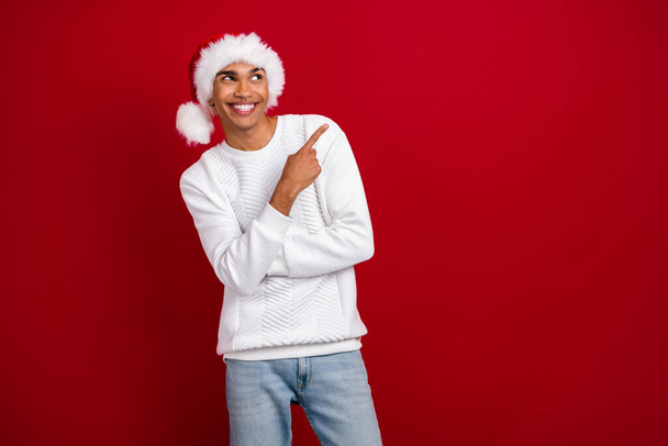 Photo of young smiling positive man wear santa claus headwear finger direct empty space look minded new winter sale isolated on red color background. - Zdjęcie, obraz