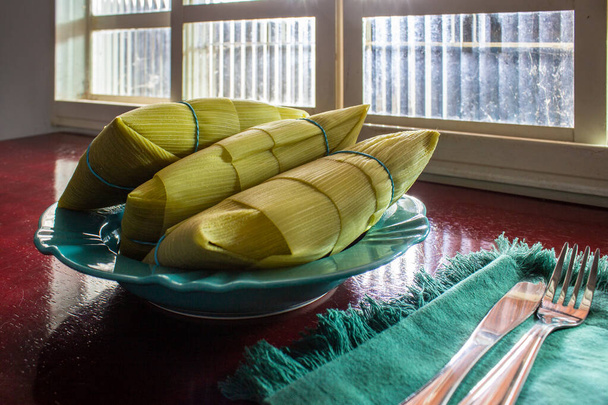 3 pamonhas covered with corn husks, inside a plate, in front of a window and on a shiny surface. - Photo, Image