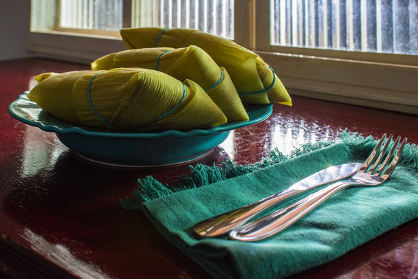 3 pamonhas cubiertas con cáscaras de maíz, dentro de un plato, frente a una ventana y sobre una superficie brillante. - Foto, Imagen