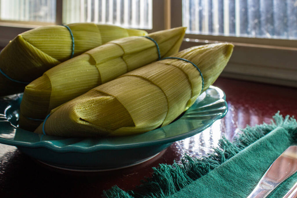 3 pamonhas cubiertas con cáscaras de maíz, dentro de un plato, frente a una ventana y sobre una superficie brillante. - Foto, Imagen