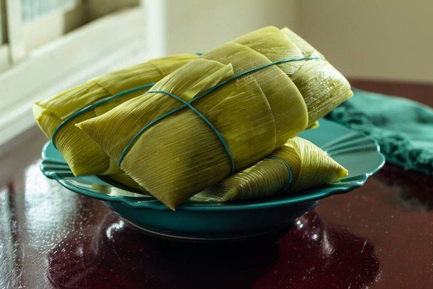 3 pamonhas cubiertas con cáscaras de maíz, dentro de un plato verde, iluminado por la luz de la ventana, sobre una superficie brillante. - Foto, Imagen