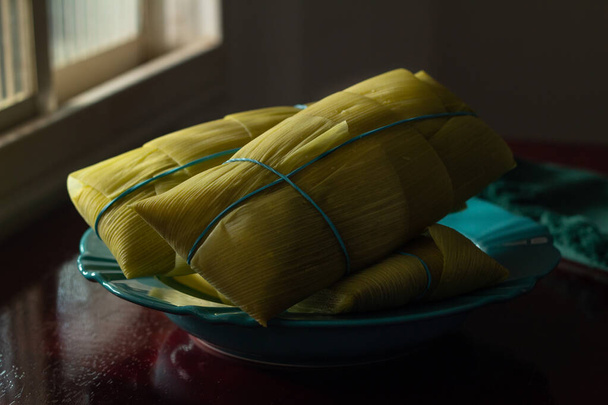 3 pamonhas cubiertas con cáscaras de maíz, dentro de un plato verde, iluminado por la luz de la ventana, sobre una superficie brillante. - Foto, imagen