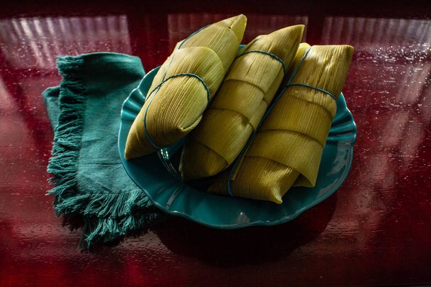 3 pamonhas cubiertas con cáscaras de maíz, dentro de un plato, sobre una superficie brillante. - Foto, imagen