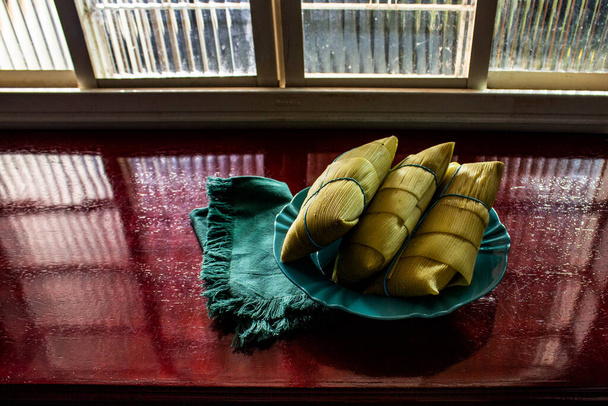 3 pamonhas recouverts d'écorces de maïs, à l'intérieur d'une assiette, devant une fenêtre et sur une surface brillante. - Photo, image