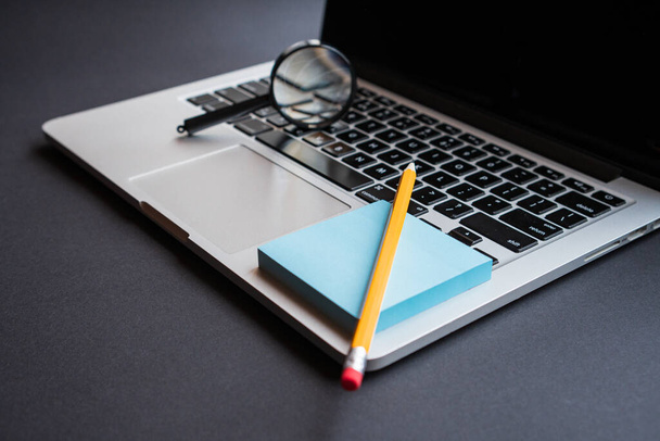 Lupe mit schwarzem Griff, gelbem Bleistift, blauem Klebezettel und grauem Laptop. Büroarbeitsplätze. - Foto, Bild