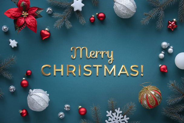 Text Merry Christmas. Festive Xmas decorations on dark turquoise emerald green textile tablecloth. Shiny golden greeting caption. Fir twigs, poinsettia flower, white and red toys, snowflakes and mirror disco balls. - Foto, imagen