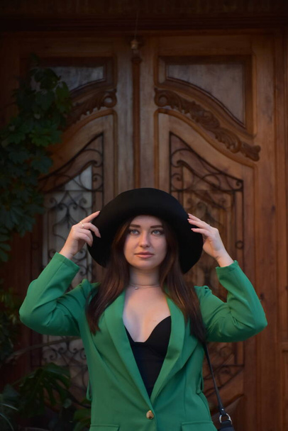 красива молода жінка в капелюсі і чорне пальто позує на фоні дерев'яного будинку
 - Фото, зображення