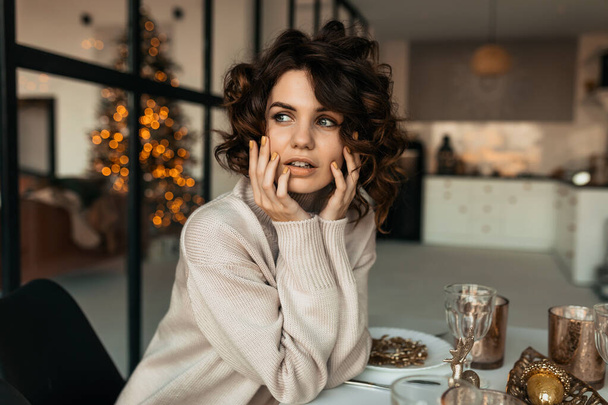 Detailní portrét elegantní stylové ženy s kudrnaté vlasy nosí svetr sedí na vánočním stolku a dívá se stranou na pozadí vánočního stromečku. - Fotografie, Obrázek