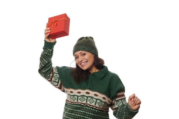 Attractive Hispanic woman in warm wool green sweater and hat, sweetly smiling looking at camera, rejoicing at Christmas present in beautiful red gift box, on white background with advertising space - Photo, Image