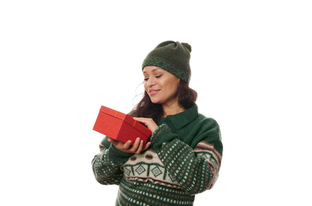 Frohe Weihnachten. Porträt einer schönen afroamerikanischen hübschen Frau in einem gemütlichen, grünen Strickpullover mit weihnachtlichem Muster und Wollmütze, die ein frohes Weihnachtsgeschenk in roter Geschenkbox hält, isoliert auf weiß - Foto, Bild