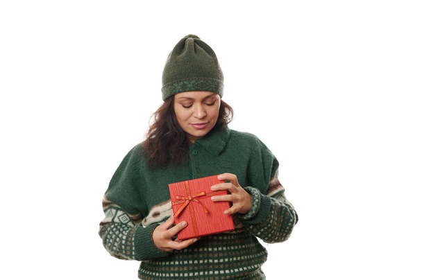 Piękna ciemnowłosa Latynoska kobieta w zielonym ciepłym wełnie Bożonarodzeniowy sweter i kapelusz, z pudełkiem świątecznego czerwonego prezentu, odizolowana na białym tle. Czas otworzyć świąteczne prezenty. Szczęśliwego Nowego Roku - Zdjęcie, obraz