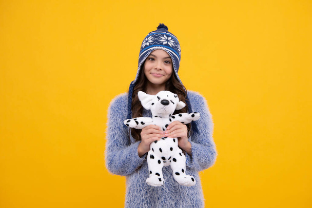 Teen girl in in winter hat hold μαλακό παιχνίδι για τα γενέθλια σε κίτρινο φόντο. Το παιδί περνάει χρόνο με τα παιχνίδια της. Παιδική ηλικία, παιχνίδια και παιδιά. Ευτυχισμένο κορίτσι. - Φωτογραφία, εικόνα