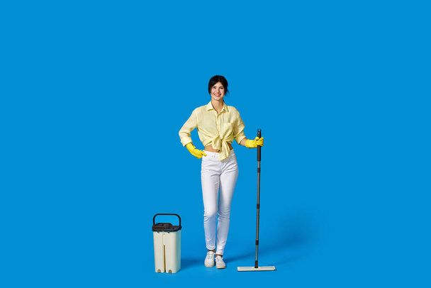 cheerful housewife woman in yellow rubber gloves washing floor with mop on blue background. Full length - Foto, Imagen