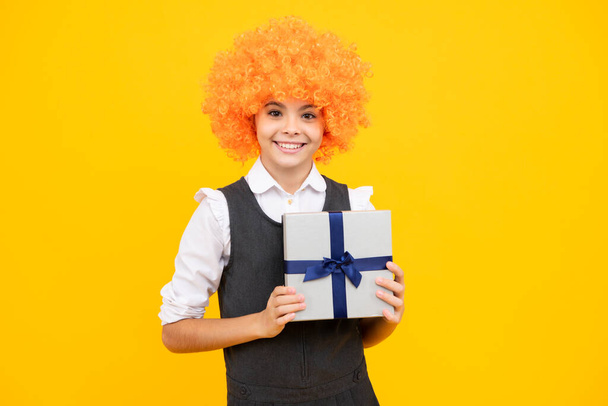 Teenager dítě v legrační paruce držení dárkové krabice na žlutém izolovaném pozadí. Dárek pro děti k narozeninám. Vánoční nebo novoroční dárková krabice. Šťastná dívka tvář, pozitivní a usměvavé emoce - Fotografie, Obrázek