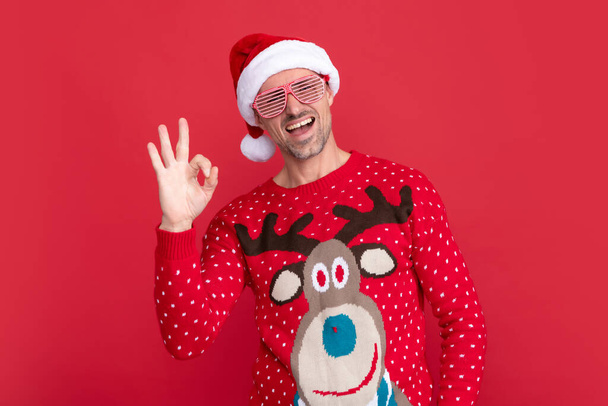 Vidám férfi téli pulcsiban és parti szemüvegben. Karácsonyi fickó télapó sapkában, piros háttérrel. Boldog új évet! Boldog karácsonyt! Rendben.. - Fotó, kép