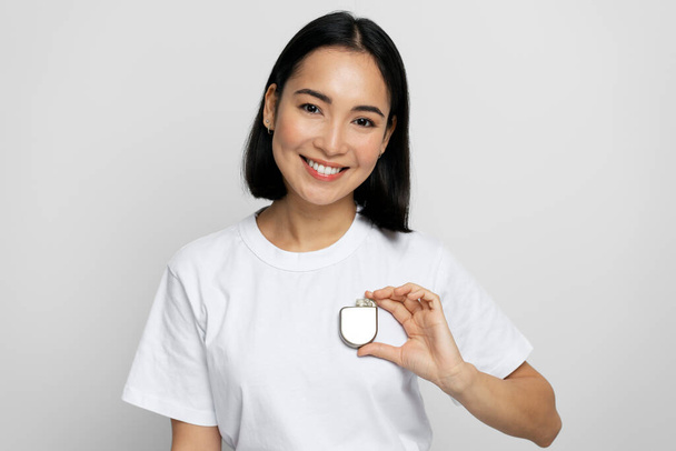 Mujer asiática positiva demostrando un desfibrilador cardioversor implantable (DAI) en las manos mientras mira a la cámara con una sonrisa agradable. Cuidado de la salud y concepto del corazón  - Foto, Imagen