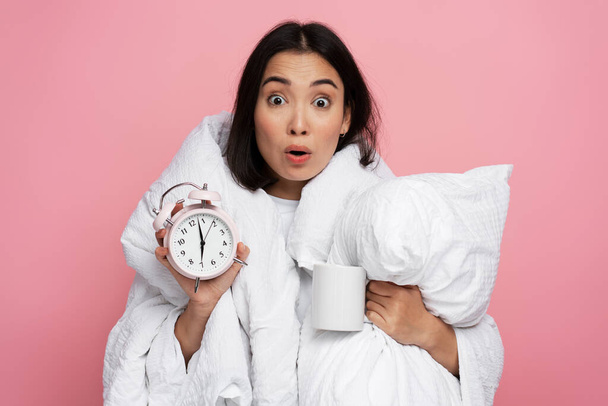 Photo portrait of surprised girl holding pillow and alarm clock with shocked face isolated on pink coloured background. Oversleep concept - Foto, afbeelding