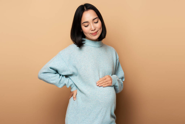 Immagine di una bella giovane donna asiatica incinta in posa isolata su sfondo beige. Emozioni felici di concetto di maternità - Foto, immagini