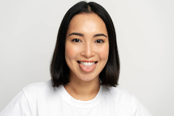 Portrait of positive playful woman standing with tongue out, demonstrating childish behaviour, wearing white T-shirt. Indoor studio shot isolated on white background  - 写真・画像