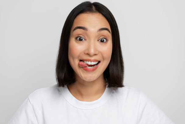 Portrait of positive playful woman standing with tongue out, demonstrating childish behaviour, wearing white T-shirt. Indoor studio shot isolated on white background  - Photo, Image