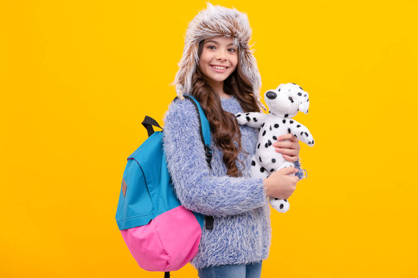 正ティーン女の子身に着けていますイヤーフラップ帽子とともに学校バッグとおもちゃで黄色の背景,バックへ学校. - 写真・画像