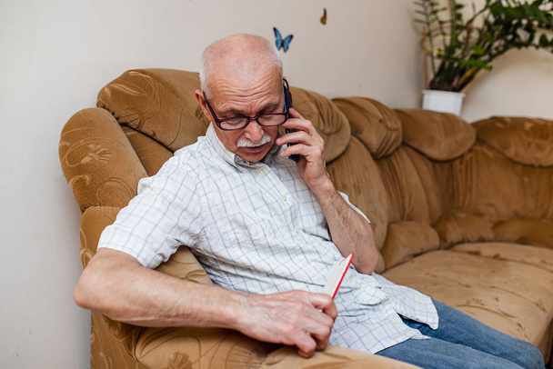 Yaşlı bir adam telefonda konuşur ve verileri bir günlüğe ya da planöre yazar. Emeklilikteki iş organizasyonu. Bir emekli uzaktan çalışır, evden iş yürütür.. - Fotoğraf, Görsel